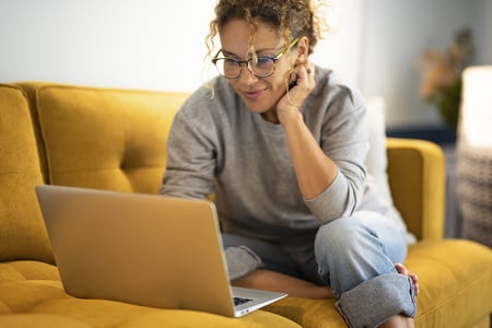 Woman on gold couch looking at real estate listings on her laptop