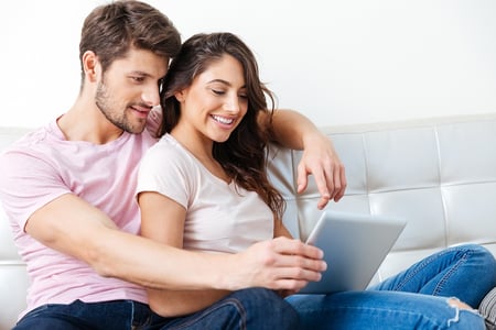 A couple sitting on a couch looking at real estate listings on a tablet. 